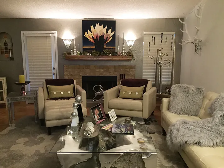 Medium-sized modern floral oil painting centered over fireplace mantel in a living room with beige armchairs and glass coffee table, demonstrating correct proportions for art above furniture